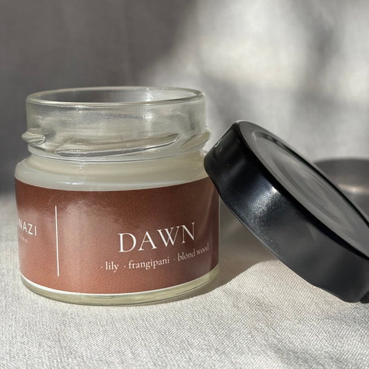 A glass jar candle with a brown label reading 'DAWN' and 'lily · frangipani · blend wood' scent, shown with its black lid resting beside it on a light fabric surface.
