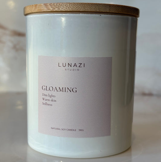 A white soy candle jar with a gold-toned metal lid and a pale pink label reading 'LUNAZI STUDIO' and 'GLOAMING'. NATURAL SOY CANDLE