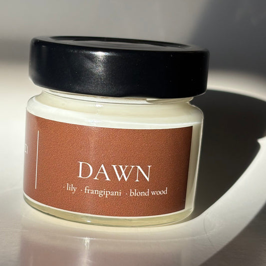 A small candle jar with a black lid and a brown label reading 'DAWN' and 'honey - frangipani - blend wood', photographed on a reflective surface with strong sunlight and shadows.