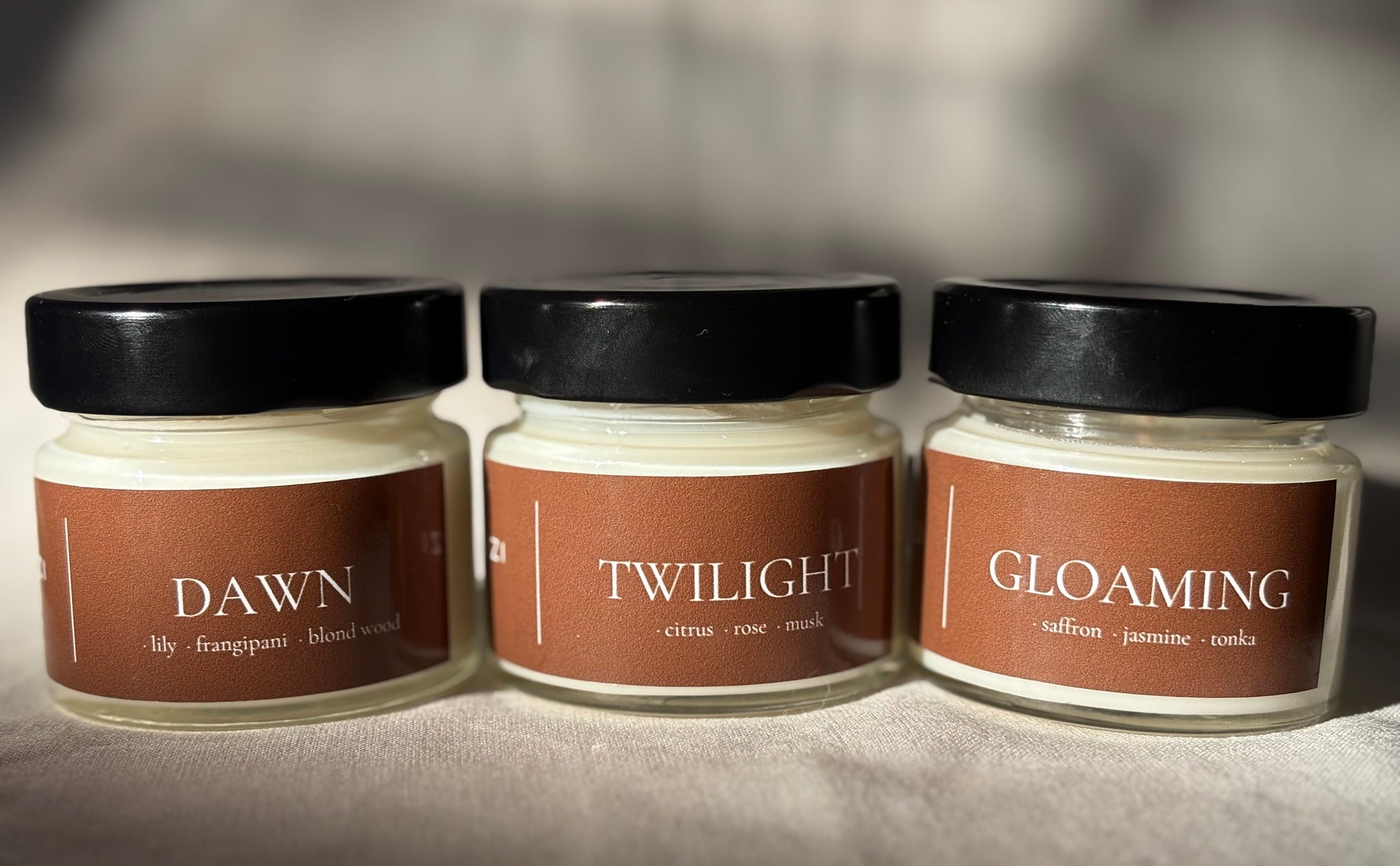 Three small glass jars of scented wax candles with black lids and brown labels reading DAWN, TWILIGHT, and GLOAMING, arranged on a fabric surface.