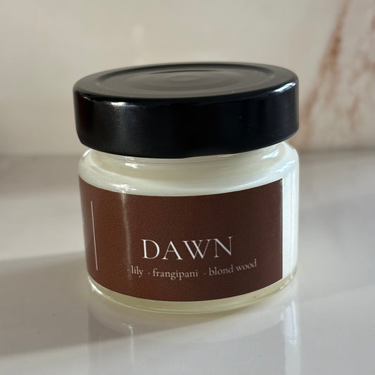 A small glass jar candle with a black lid and a brown label reading 'DAWN' and 'lily - frangipani - blond wood', photographed on a light surface with a soft neutral background.