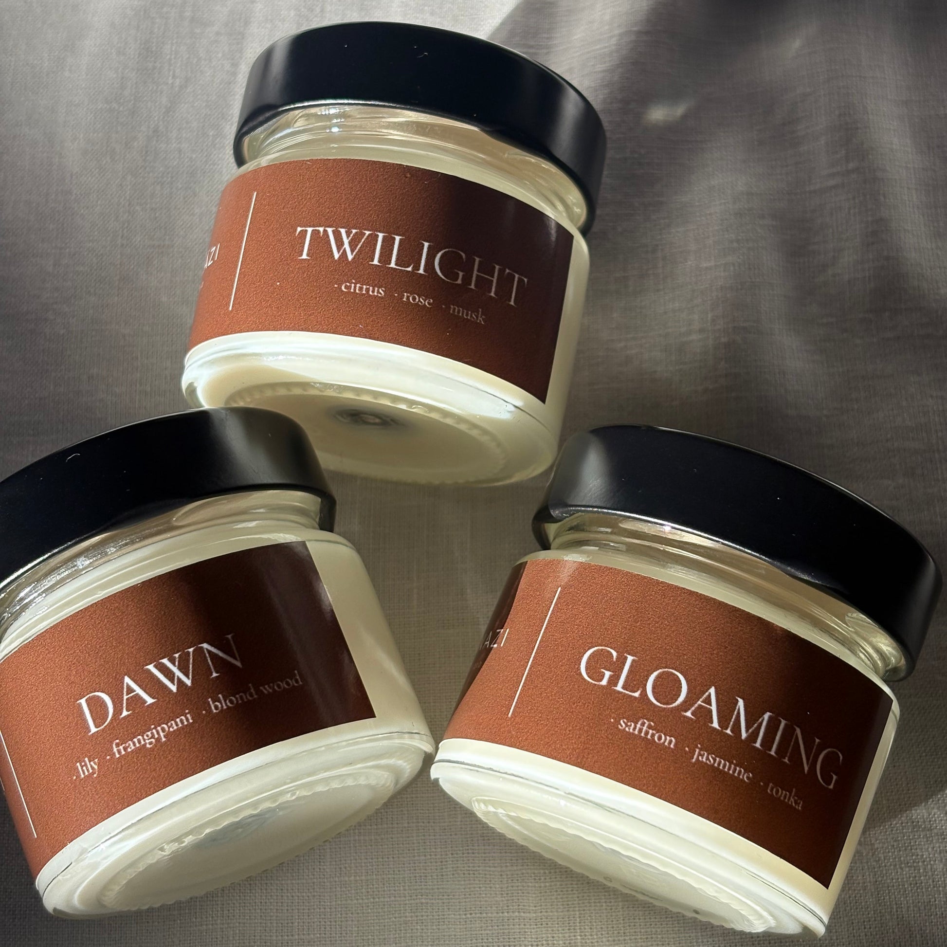 Three glass jars of scented candles with black lids and brown labels, labeled Twilight, Gloaming, and Dawn, arranged on a gray fabric background.