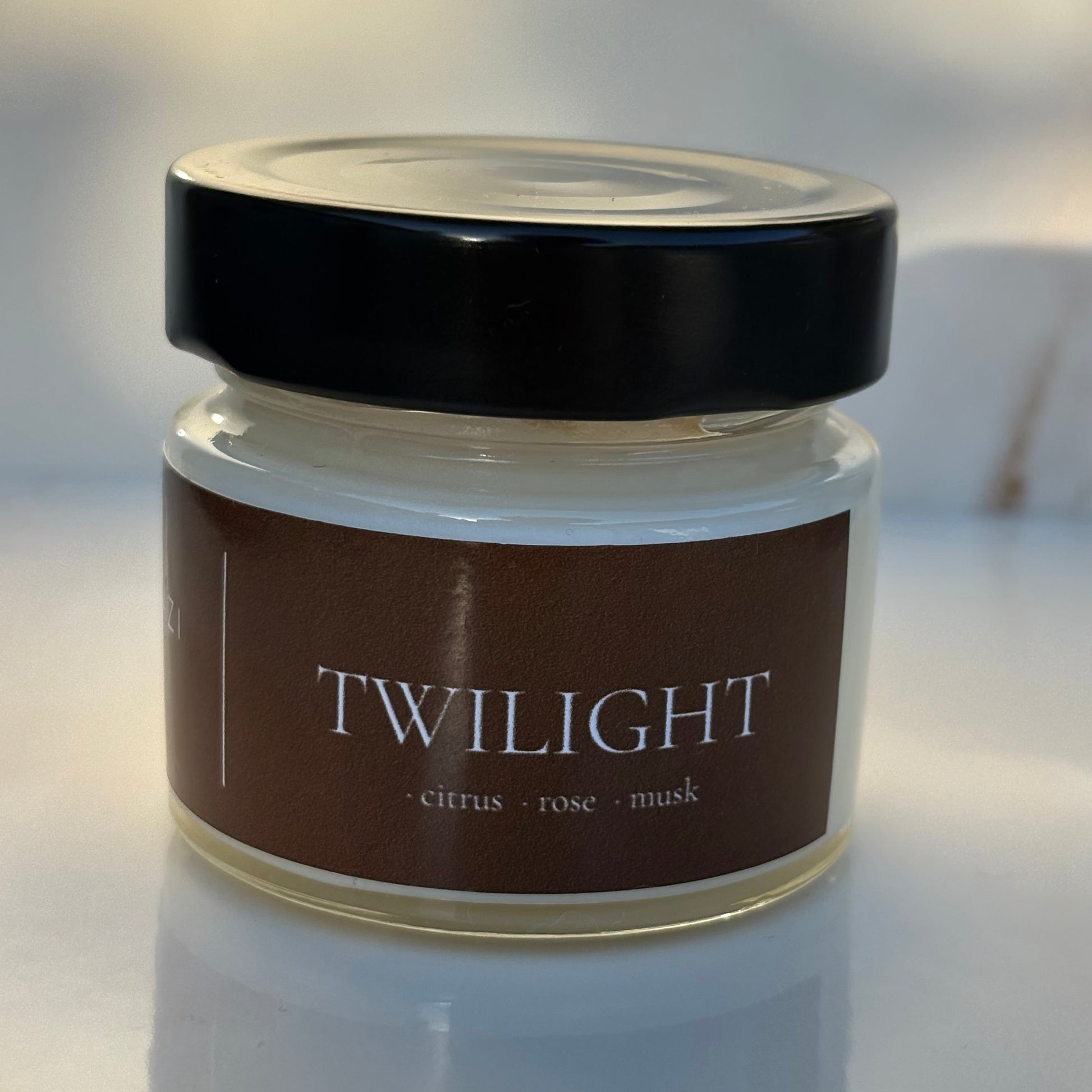 A small glass jar candle with a black lid and a dark brown label reading 'TWILIGHT' and 'citrus • rose • musk', photographed on a light reflective surface.
