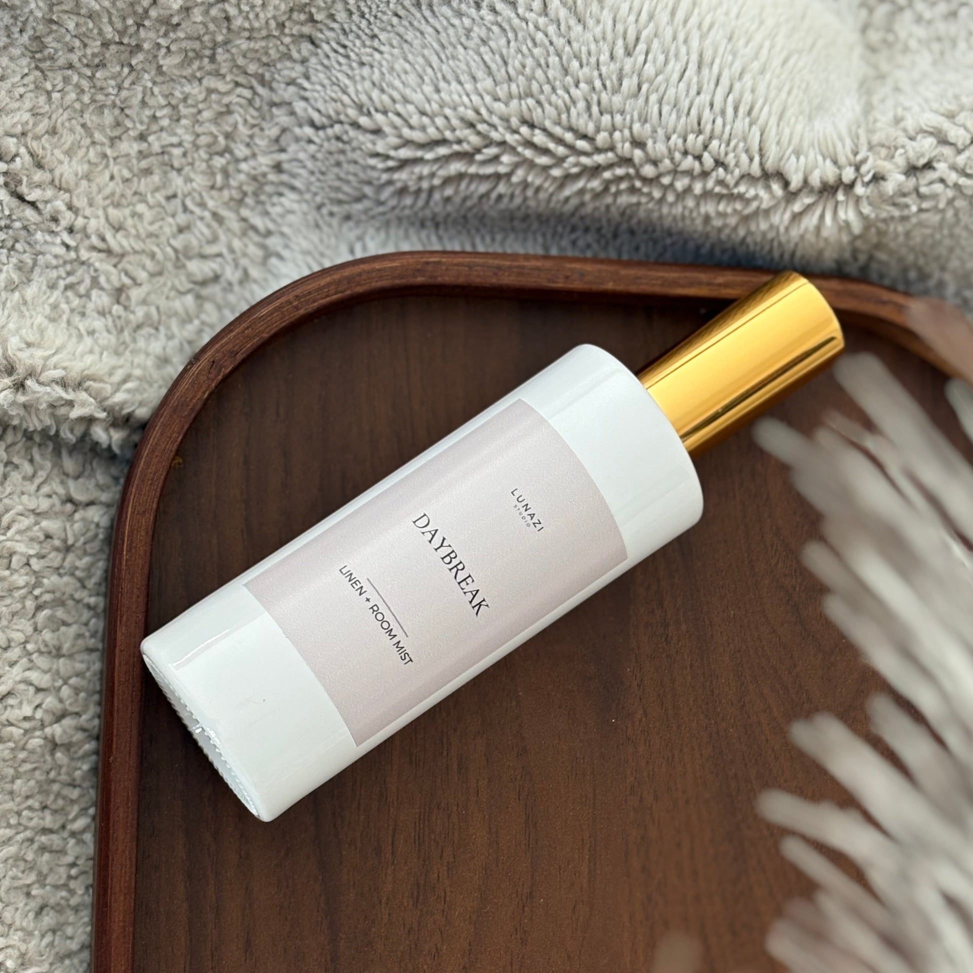 A bottle of Lunazi Daybreak Linen + Room Mist, featuring a white cylindrical bottle with a light gray label and a dark brown cap, set on a brown tray surface with a warm beige background.