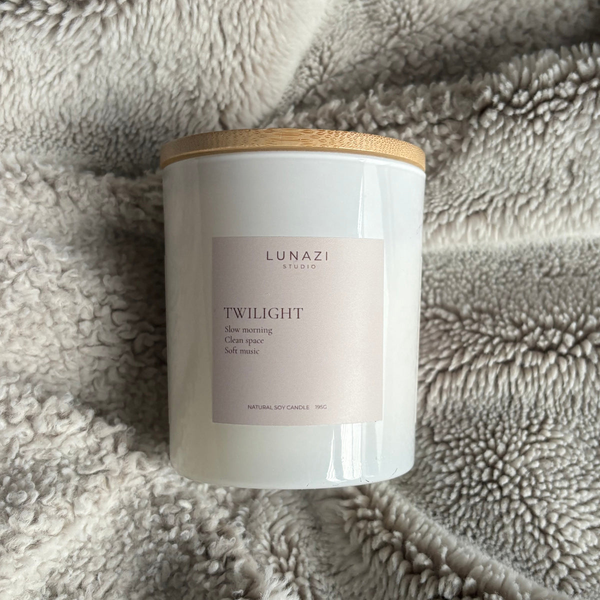 A white jar candle with a light wood lid and a pale pink label reading 'LUNAZI STUDIO TWILIGHT scented oil candle,' placed on a soft gray textured blanket.