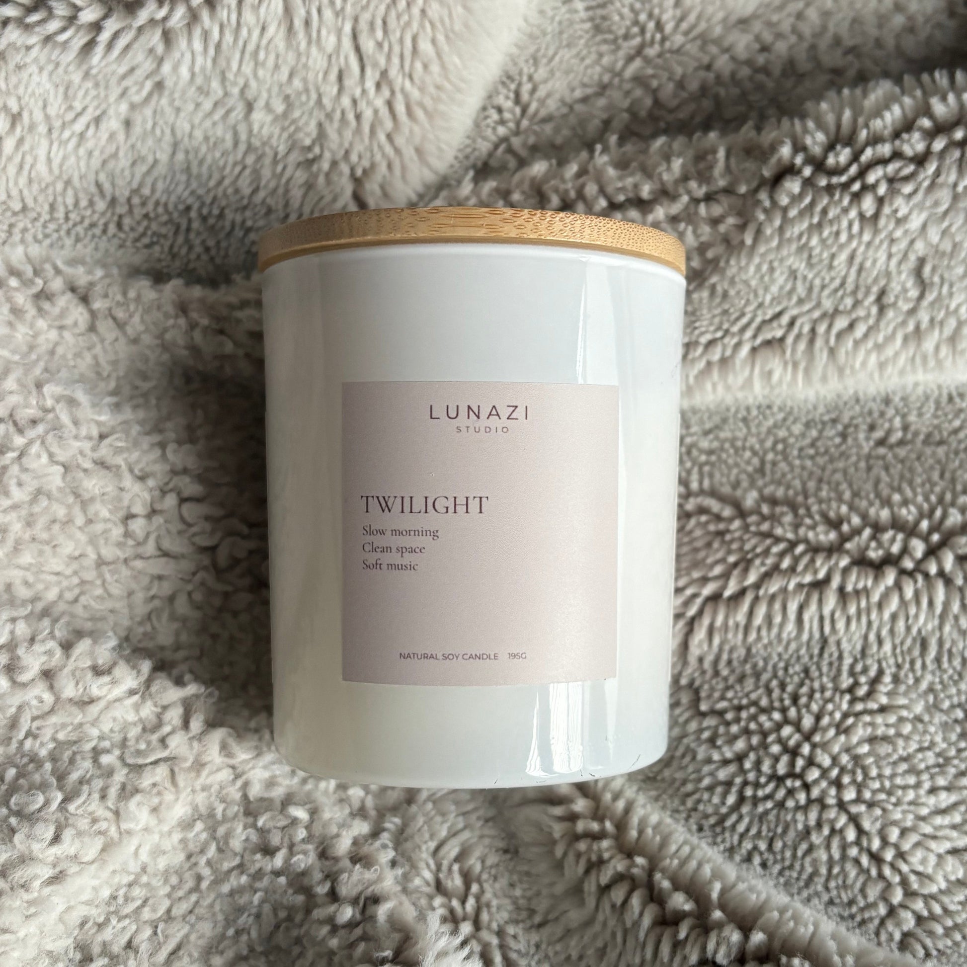 A white jar candle with a light wood lid and a pale pink label reading 'LUNAZI STUDIO TWILIGHT scented oil candle,' placed on a soft gray textured blanket.