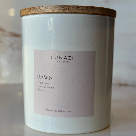 A white glass candle jar with a light wood lid, featuring a pale pink label that reads 'LUNAZI STUDIO' and 'DAWN'.