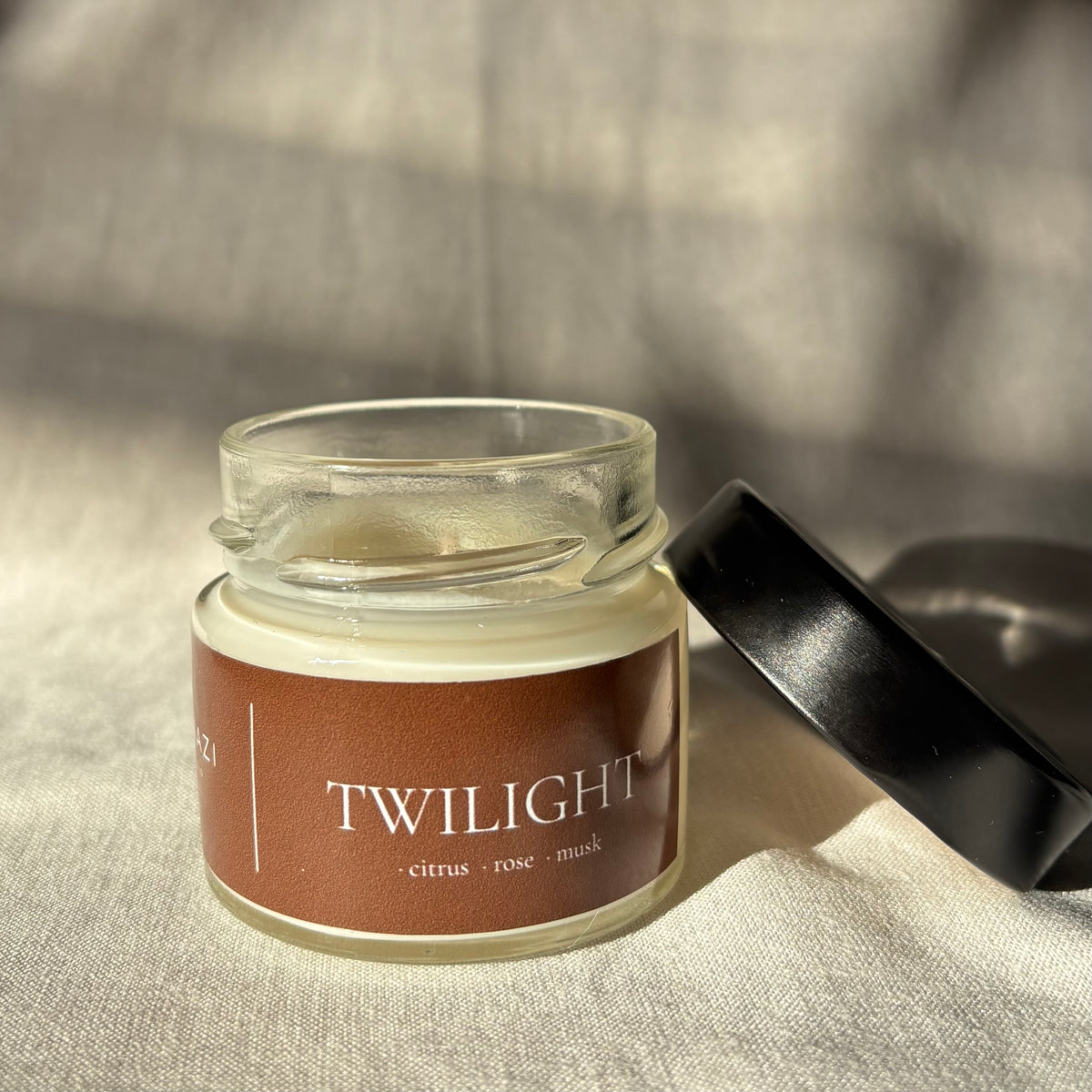A small clear glass jar candle with a brown label reading 'TWILIGHT - citrus - rose - musk' and a black lid resting beside it on a textured beige fabric background.