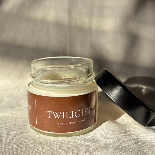A small clear glass jar candle with a brown label reading 'TWILIGHT - citrus - rose - musk' and a black lid resting beside it on a textured beige fabric background.