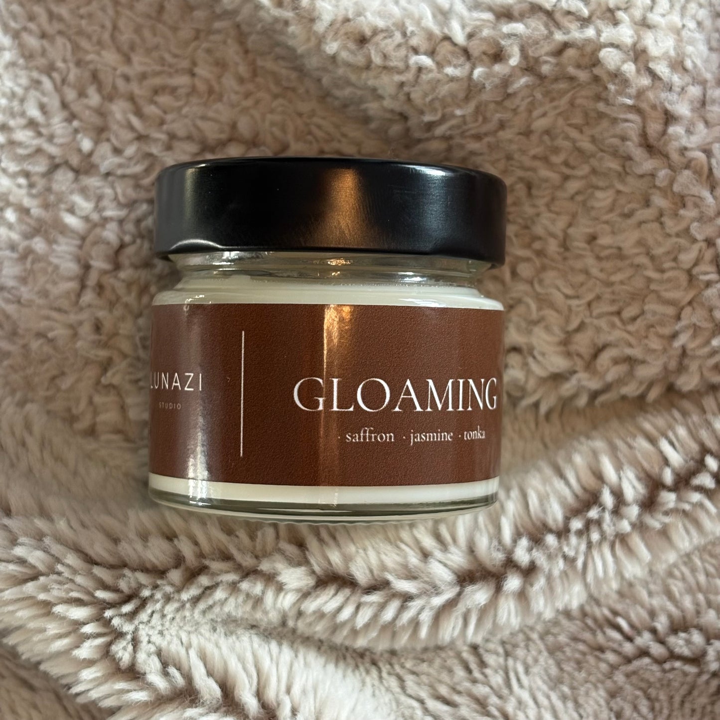 A glass jar candle with a black lid and a brown label reading 'GLOAMING' and 'saffron jasmine tea tree', placed on a beige textured fleece blanket.