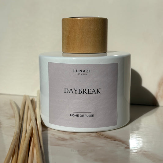 A Lunazi Studios Daybreak home diffuser with a white ceramic body, a light wood cap, and a lavender-gray label, shown on a marble surface with reed sticks in the foreground.