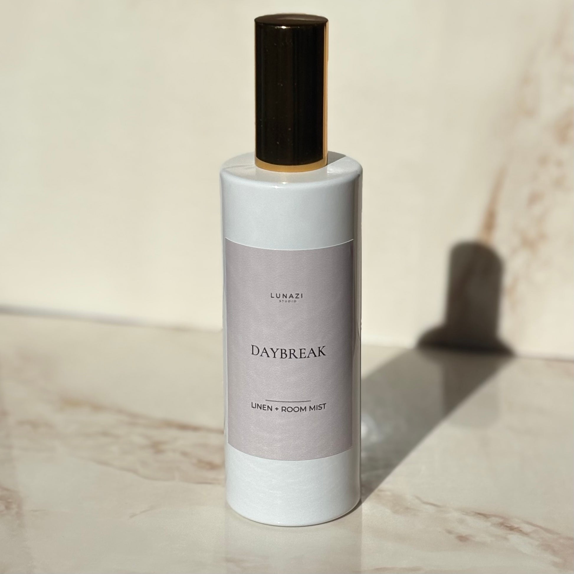 A bottle of Lunazi Daybreak Linen + Room Mist with a white body, a muted lavender label, and a dark brown cap, set on a light marble surface with a soft shadow.