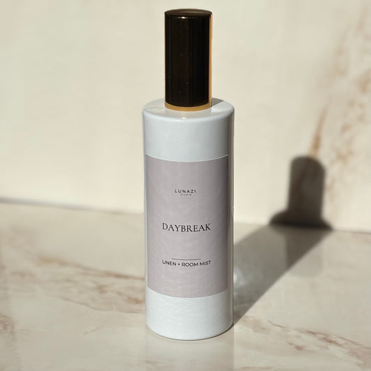 A bottle of Lunazi Daybreak Linen + Room Mist with a white body, a muted lavender label, and a dark brown cap, set on a light marble surface with a soft shadow.