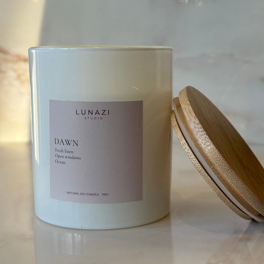 A white glass jar candle with a light lavender label reading 'LUNAZI DAWN' and a bamboo lid resting diagonally on a light marble surface.