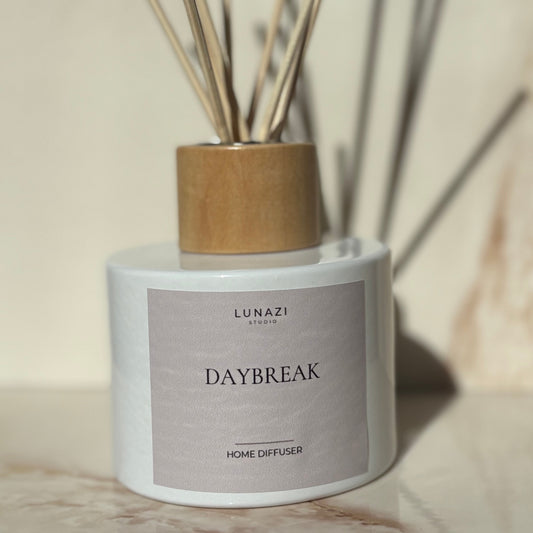 A Lunazi Daybreak home diffuser with a white cylindrical bottle, a light wood cap, and natural reed sticks, set on a beige marble surface with a matching marble background.