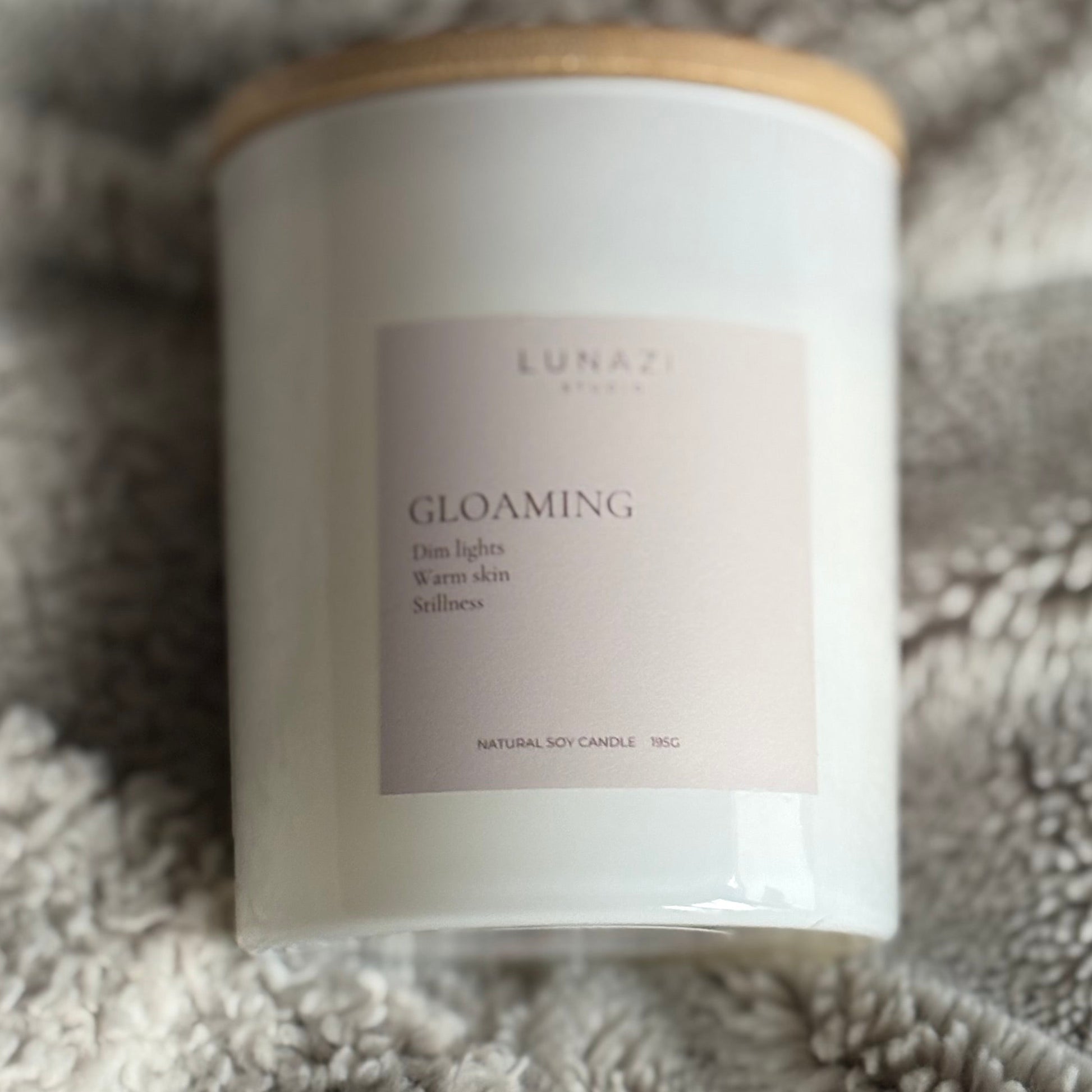 A white candle jar with a light wood lid and a pale blush label reading 'GLOAMING' and 'LUNAZI STUDIO', placed on a soft gray fleece blanket.
