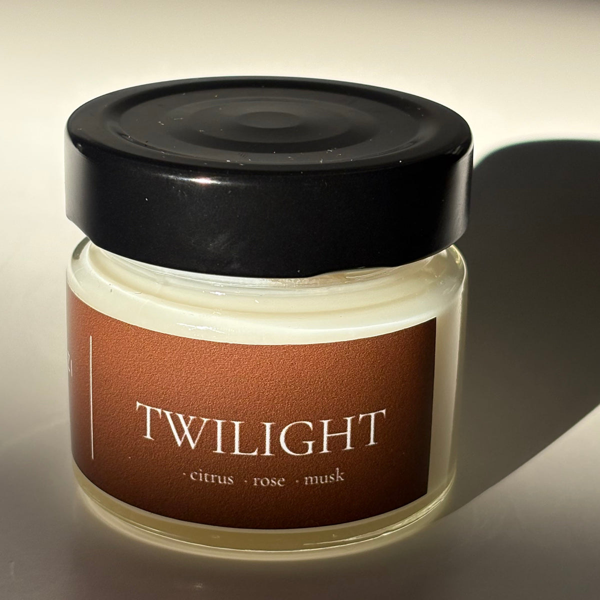 A small cosmetic jar with a black lid and a warm brown label reading 'TWILIGHT' in white text, photographed against a light neutral background with strong directional shadows.