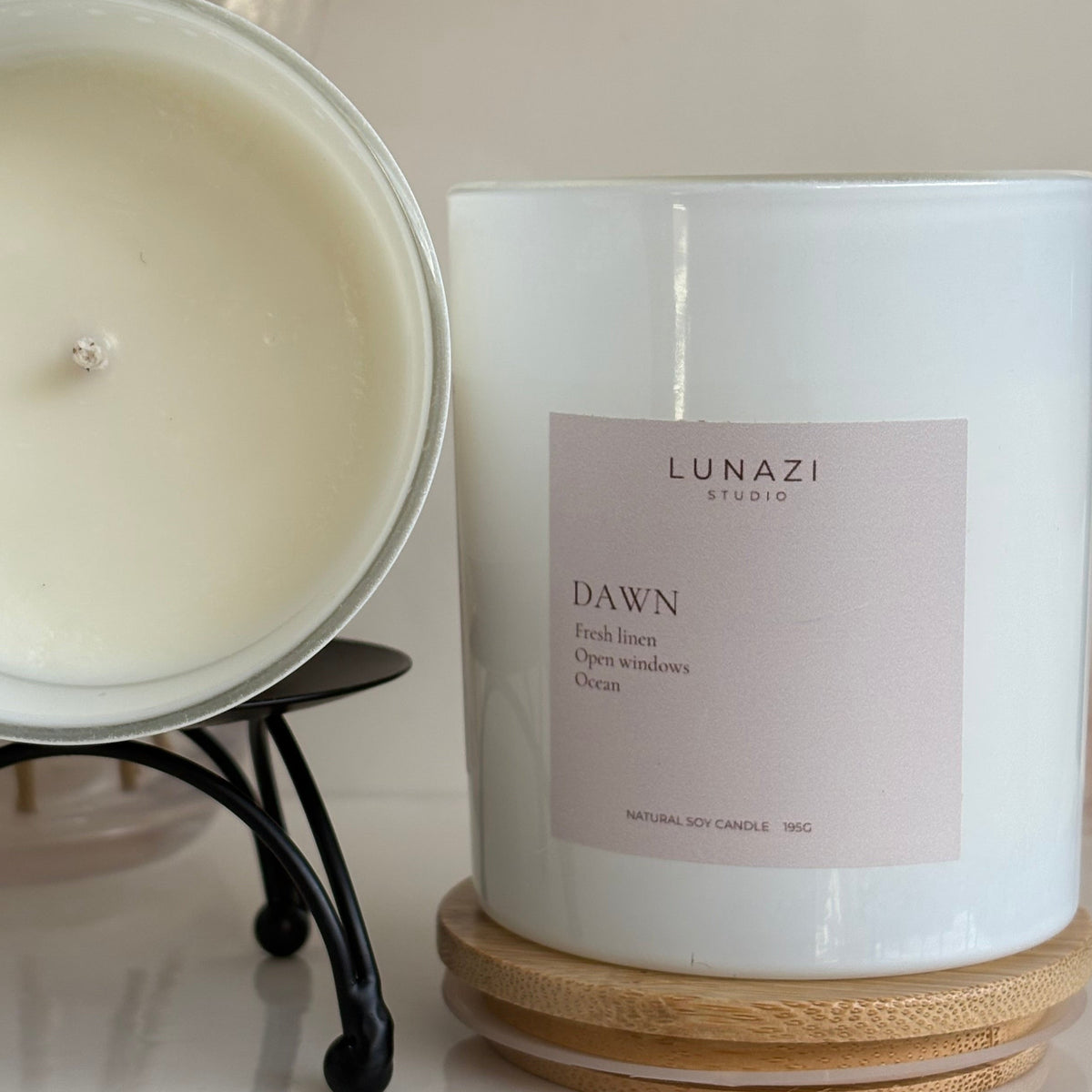 A white soy candle shown in two views: one sliced open on a black stand to reveal the creamy off-white wax, and one whole candle in a glass jar with a pale pink label reading 'LUNAZI STUDIO DAWN'. The whole candle sits on a wooden coaster on a light surface, with a softly blurred decorative background.