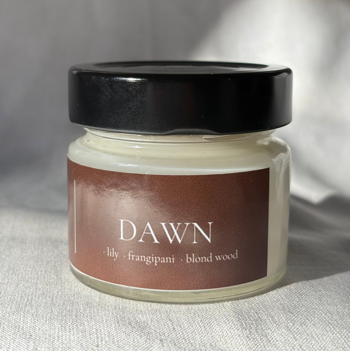A glass jar candle with creamy off-white wax, a black lid, and a brown label reading 'DAWN' with scent notes 'lily • frangipani • blond wood', photographed on a light fabric surface.