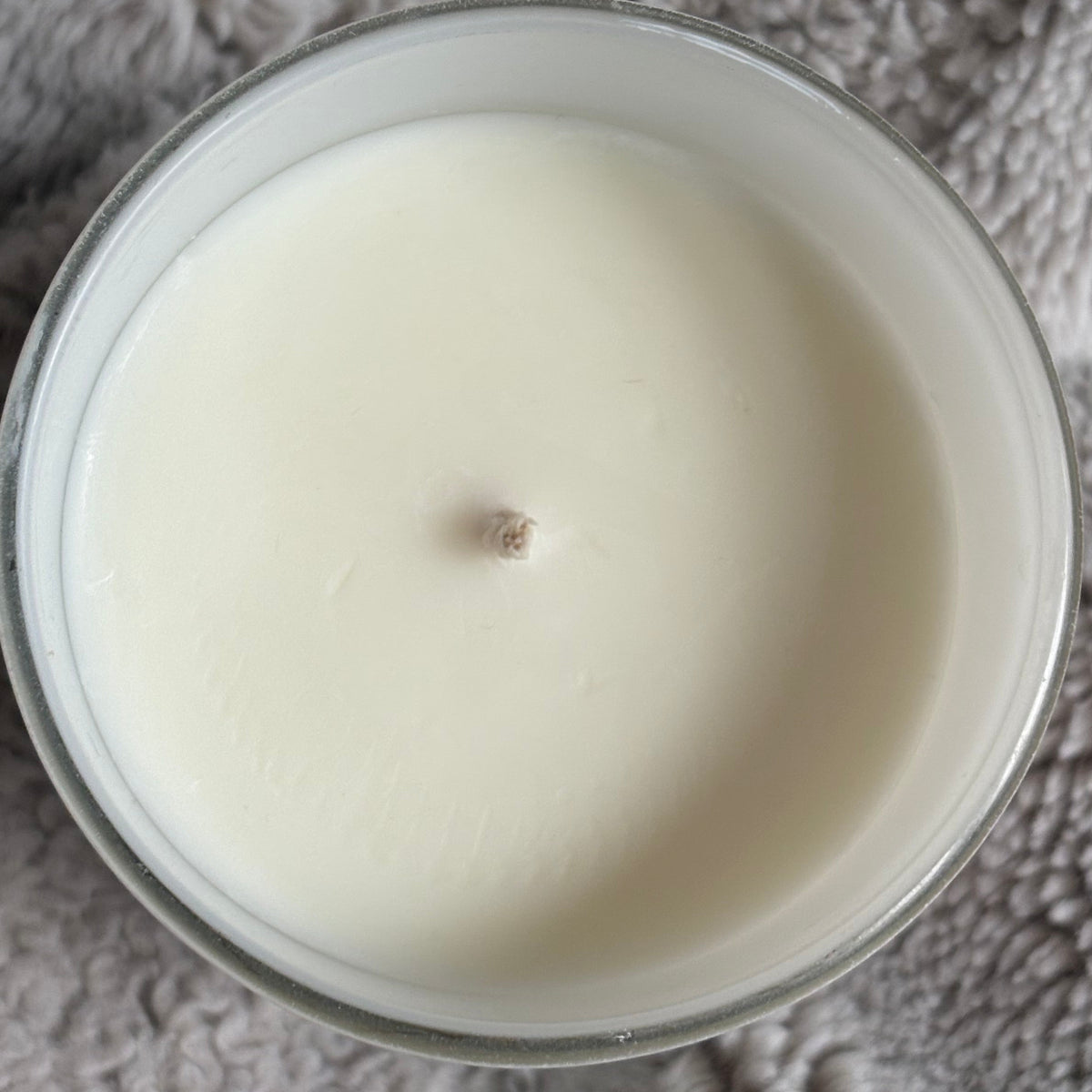 A top-down view of a white wax candle in a white glass jar, resting on a textured gray 
blanket. The image displays a cotton wick, flat and even top view of a luxury scented candle. 