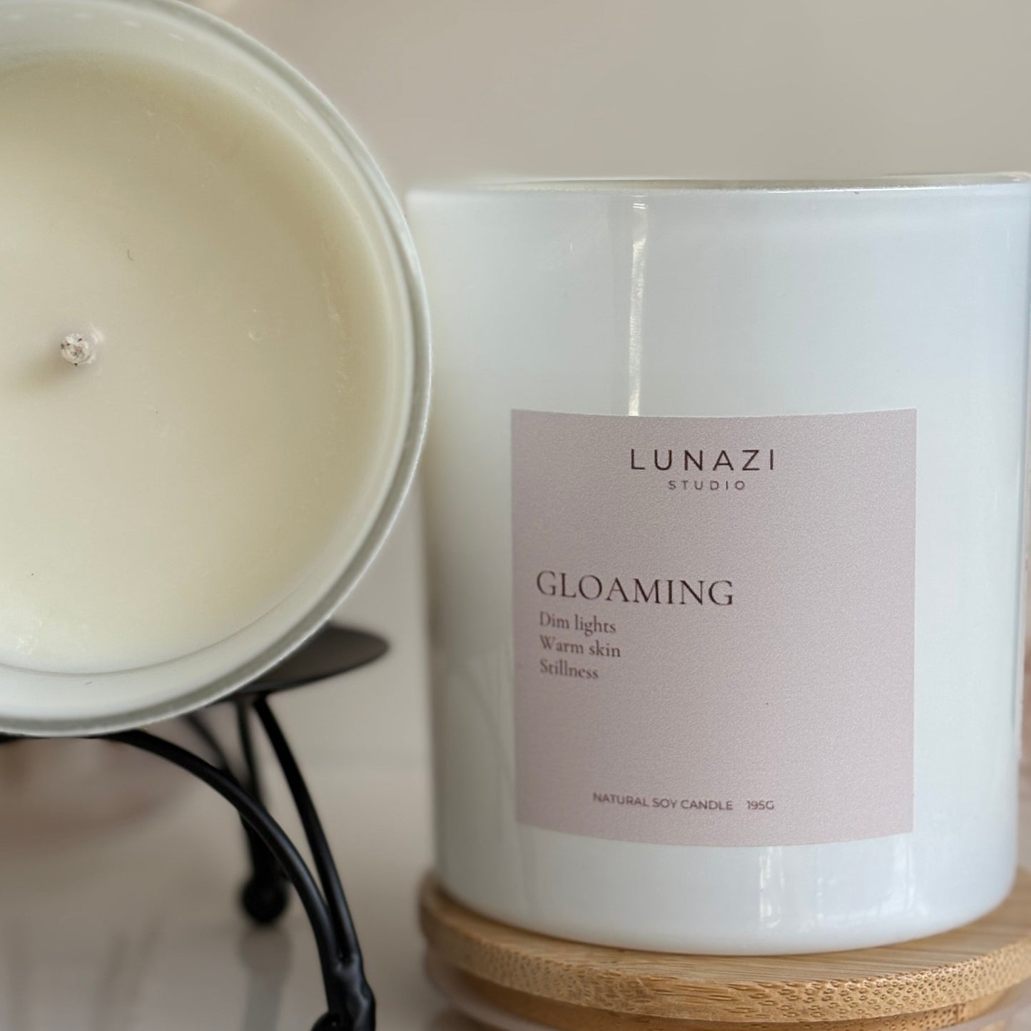 A glossy white soy candle jar labeled 'LUNAZI STUDIO GLOAMING' sits next to a second candle shown open on a small black stand, with a light wood coaster underneath. The background is a soft, warm beige with blurred dried botanicals.