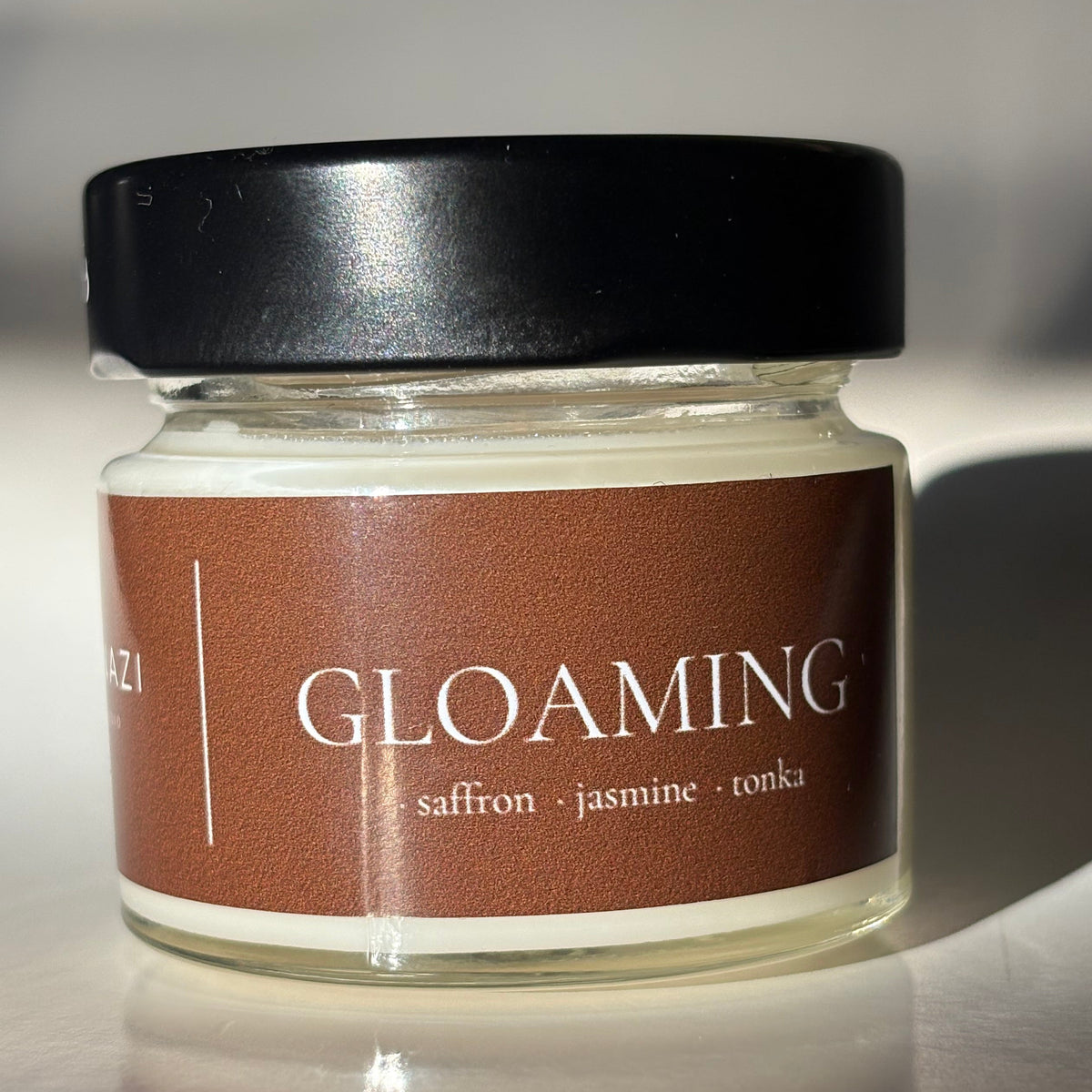 A glass jar candle with a black lid and a brown label reading 'GLOAMING' and 'saffron + jasmine + oakmoss', photographed on a light surface with strong shadows.