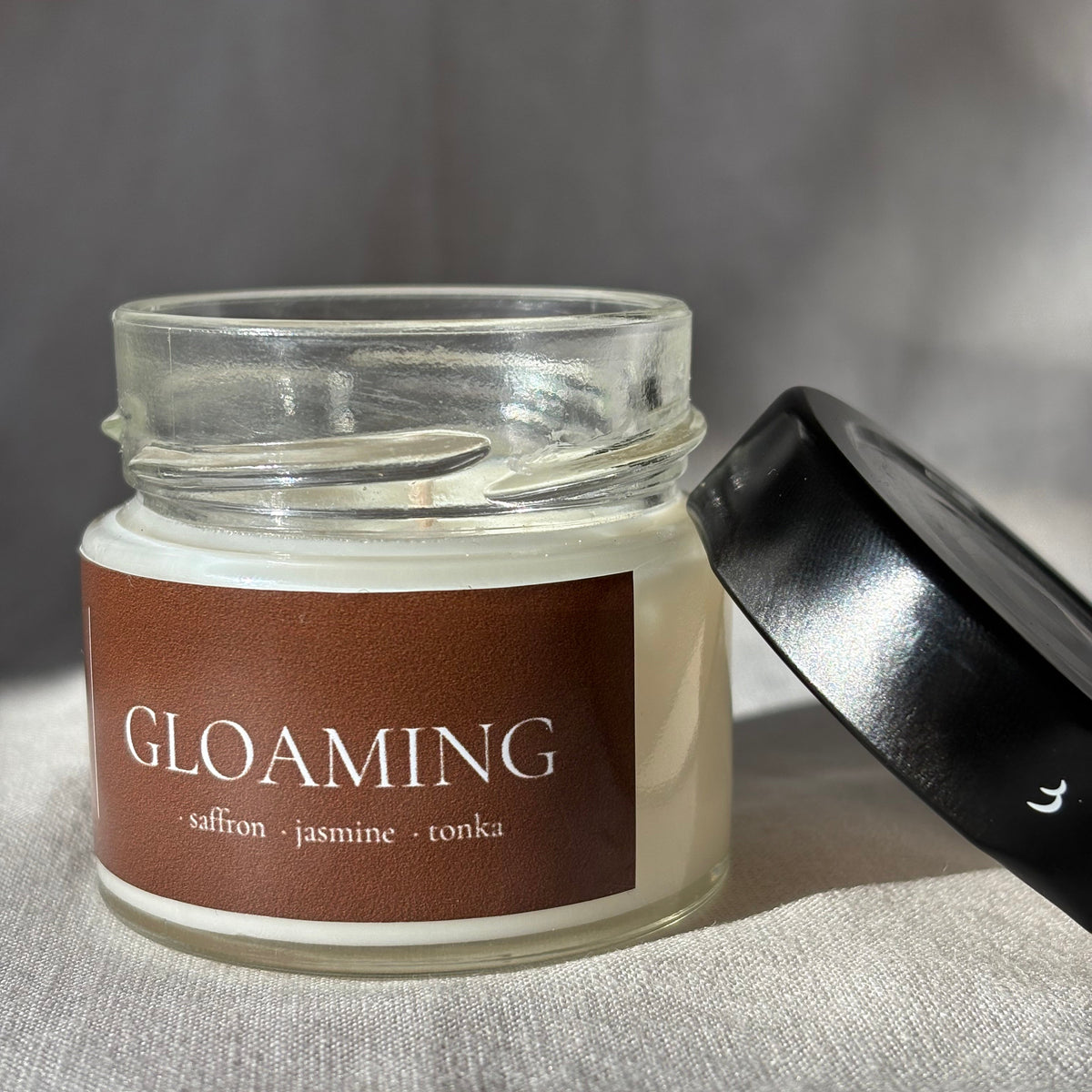 A glass jar candle with creamy off-white wax and a brown label reading 'GLOAMING' with smaller scent notes, shown with its black lid resting beside it on a textured fabric surface.