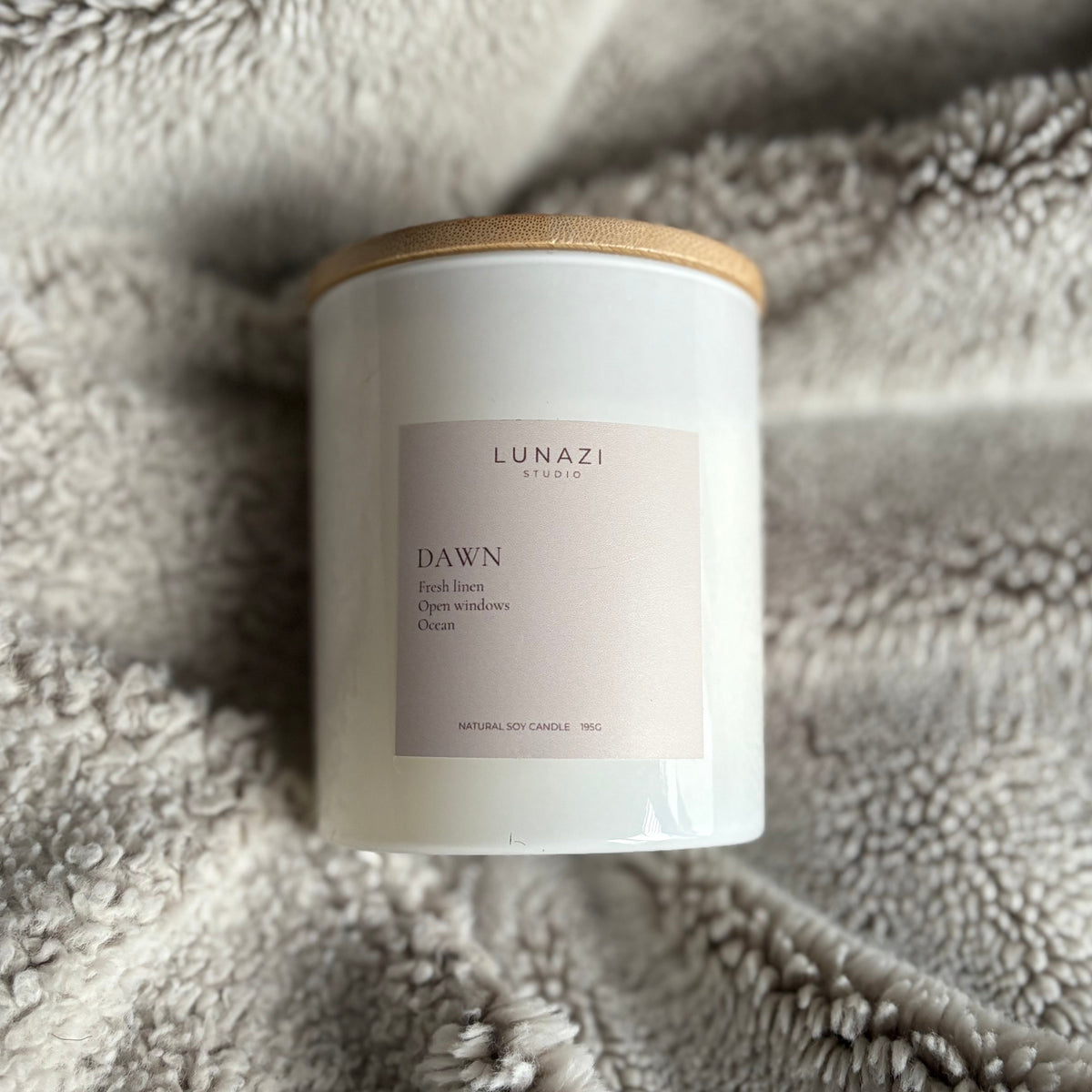 A white jar candle with a light wood lid and a pale pink label reading 'LUNAZI STUDIO DAWN scented oil candle,' placed on a soft gray textured blanket.