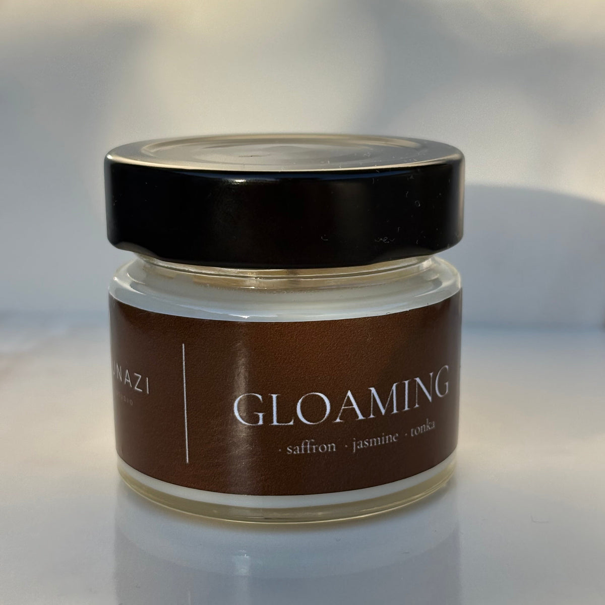 A small glass jar of 'Gloaming' body butter or cream with a dark brown label and a black lid, set against a softly blurred light gray background.