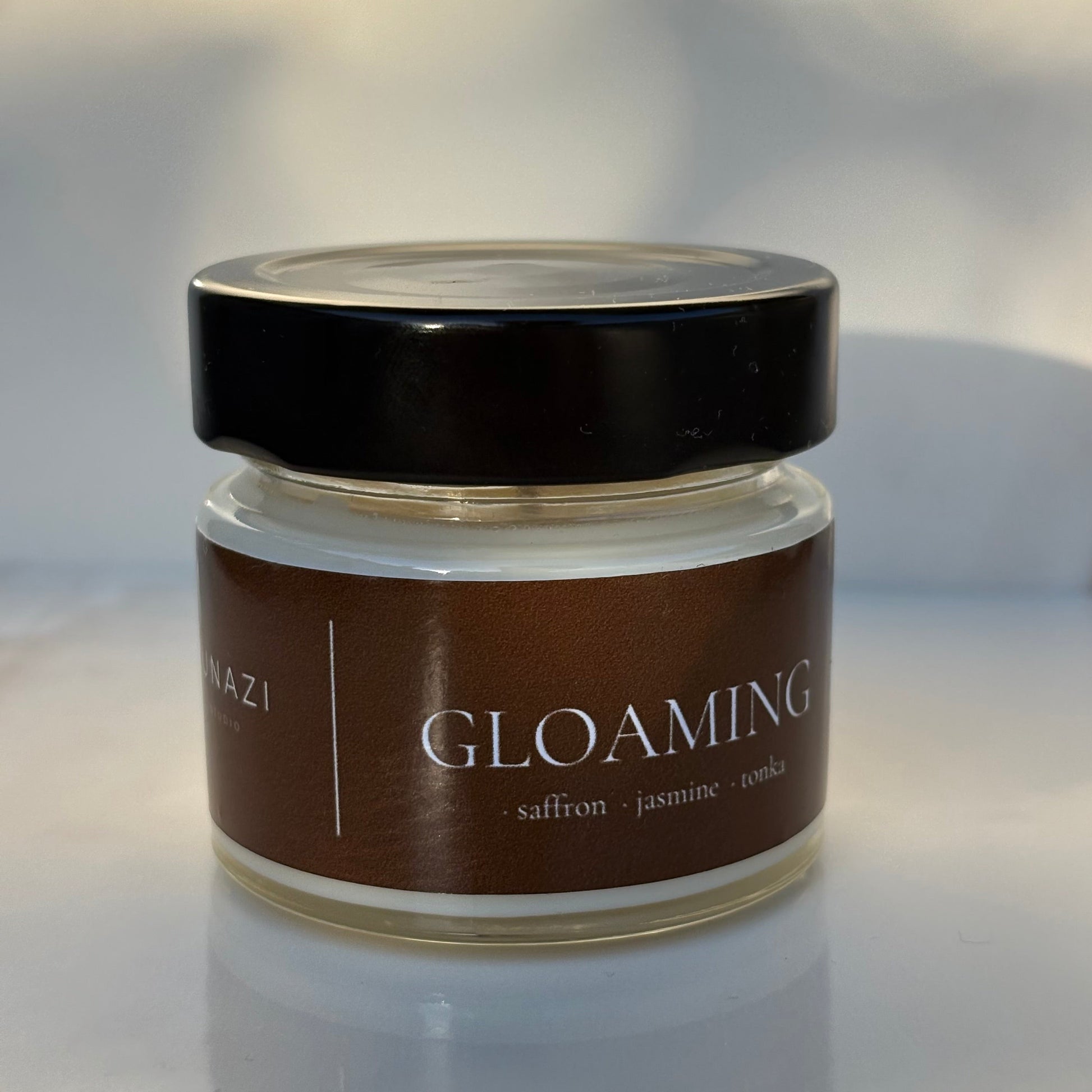A small glass jar of 'Gloaming' body butter or cream with a dark brown label and a black lid, set against a softly blurred light gray background.
