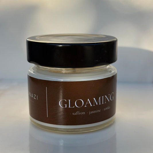 A small glass jar of 'Gloaming' body butter or cream with a dark brown label and a black lid, set against a softly blurred light gray background.