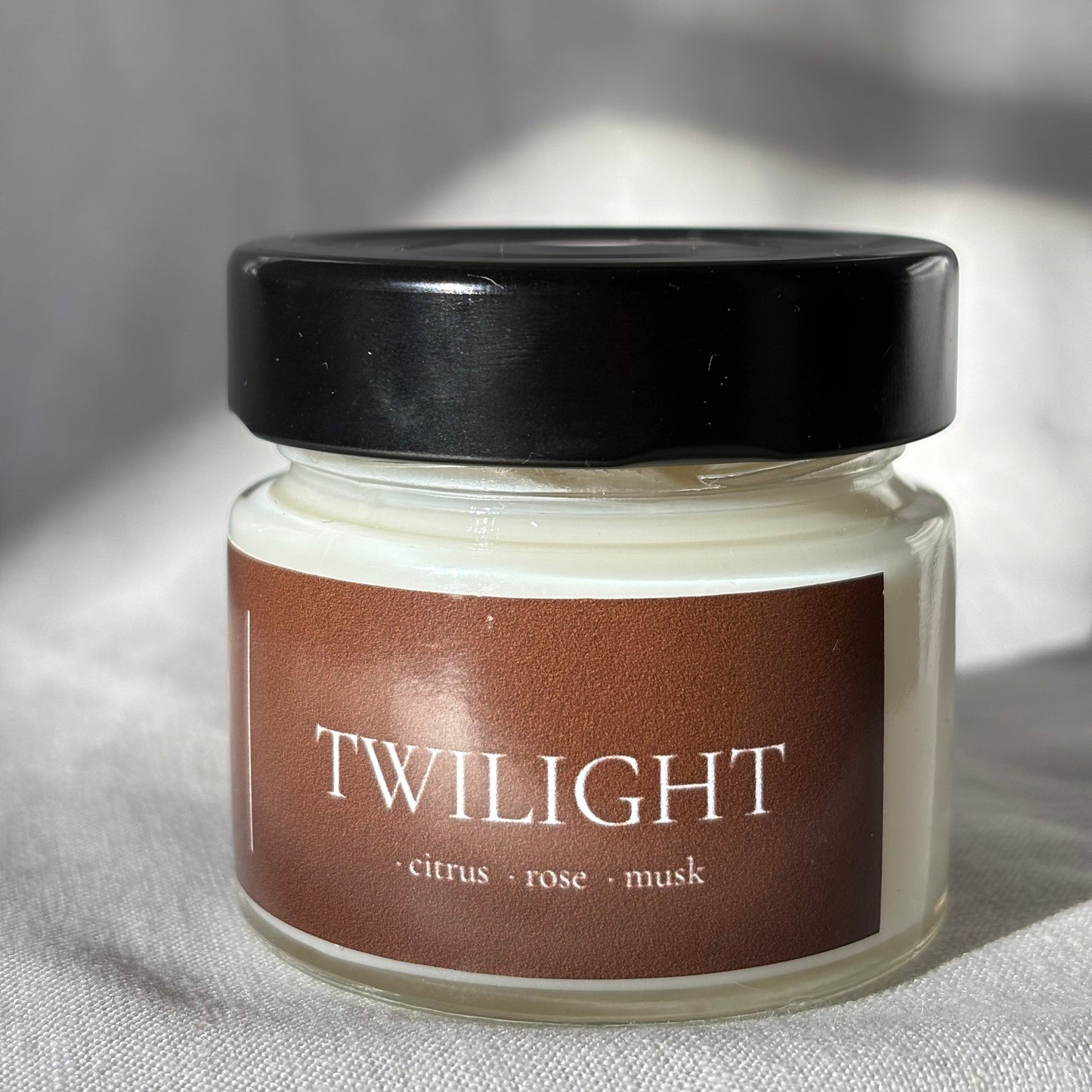 A glass jar of face mask with a black lid and a brown label reading 'TWILIGHT citrus + rose + mask', photographed on a light fabric background.