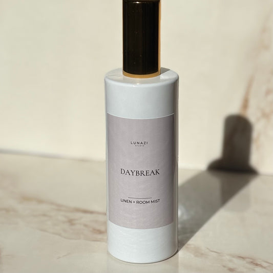 A bottle of Lunazi Daybreak Linen + Room Mist, featuring a white cylindrical bottle with a light gray label and a dark brown cap, set on a marble surface with a warm beige background.