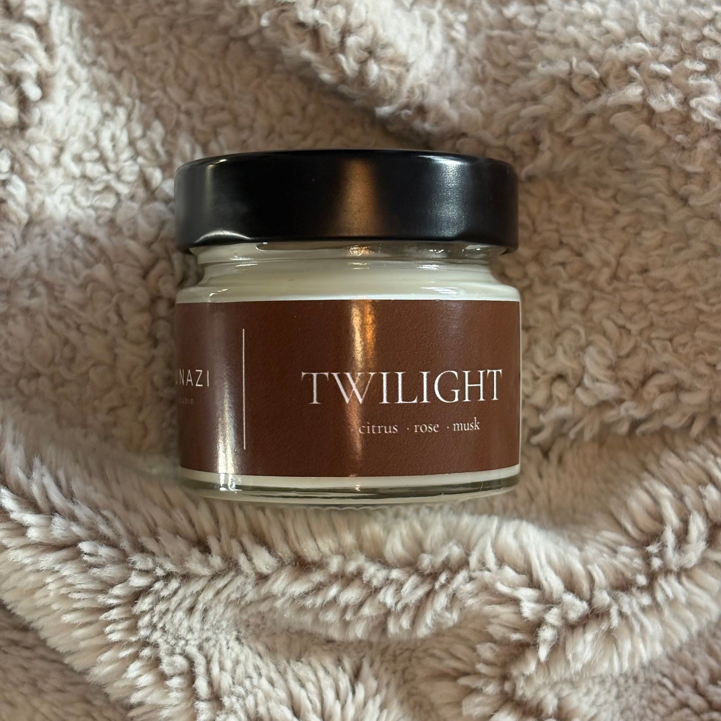 A glass jar candle with a black lid and a brown label reading 'TWILIGHT' and 'citrus + rose + mask', placed on a soft beige textured blanket.