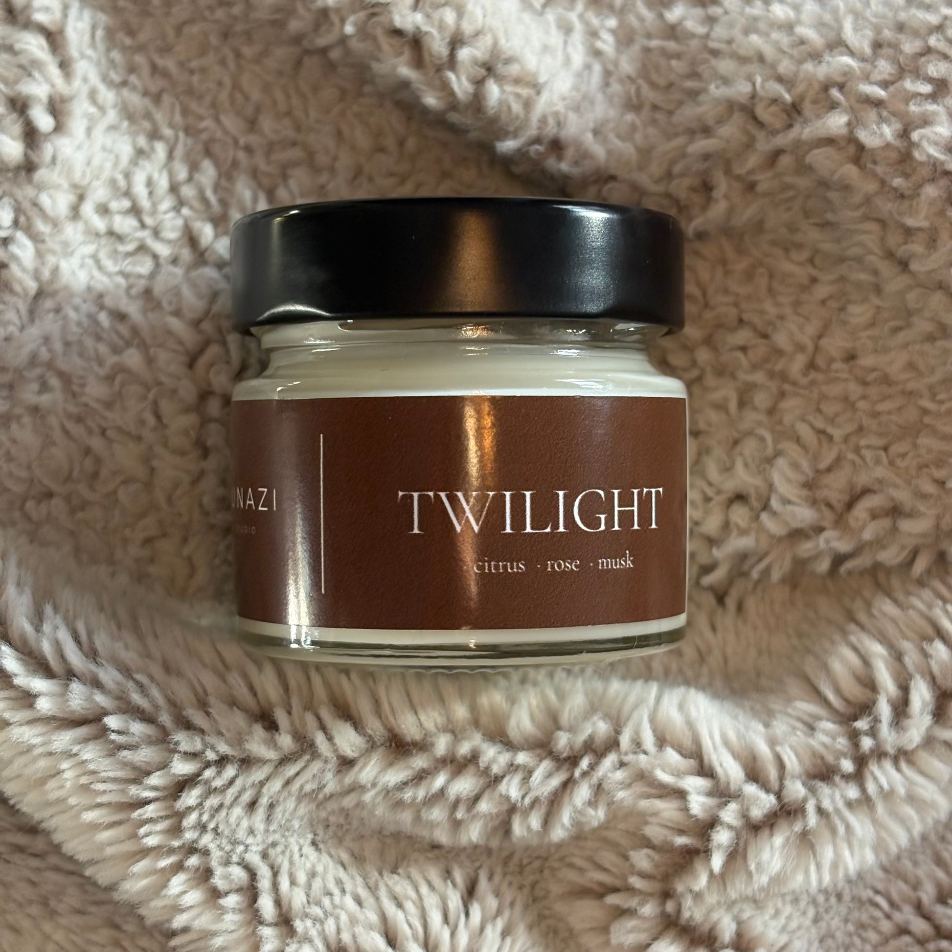 A glass jar candle with a black lid and a brown label reading 'TWILIGHT' and 'citrus + rose + mask', placed on a soft beige textured blanket.