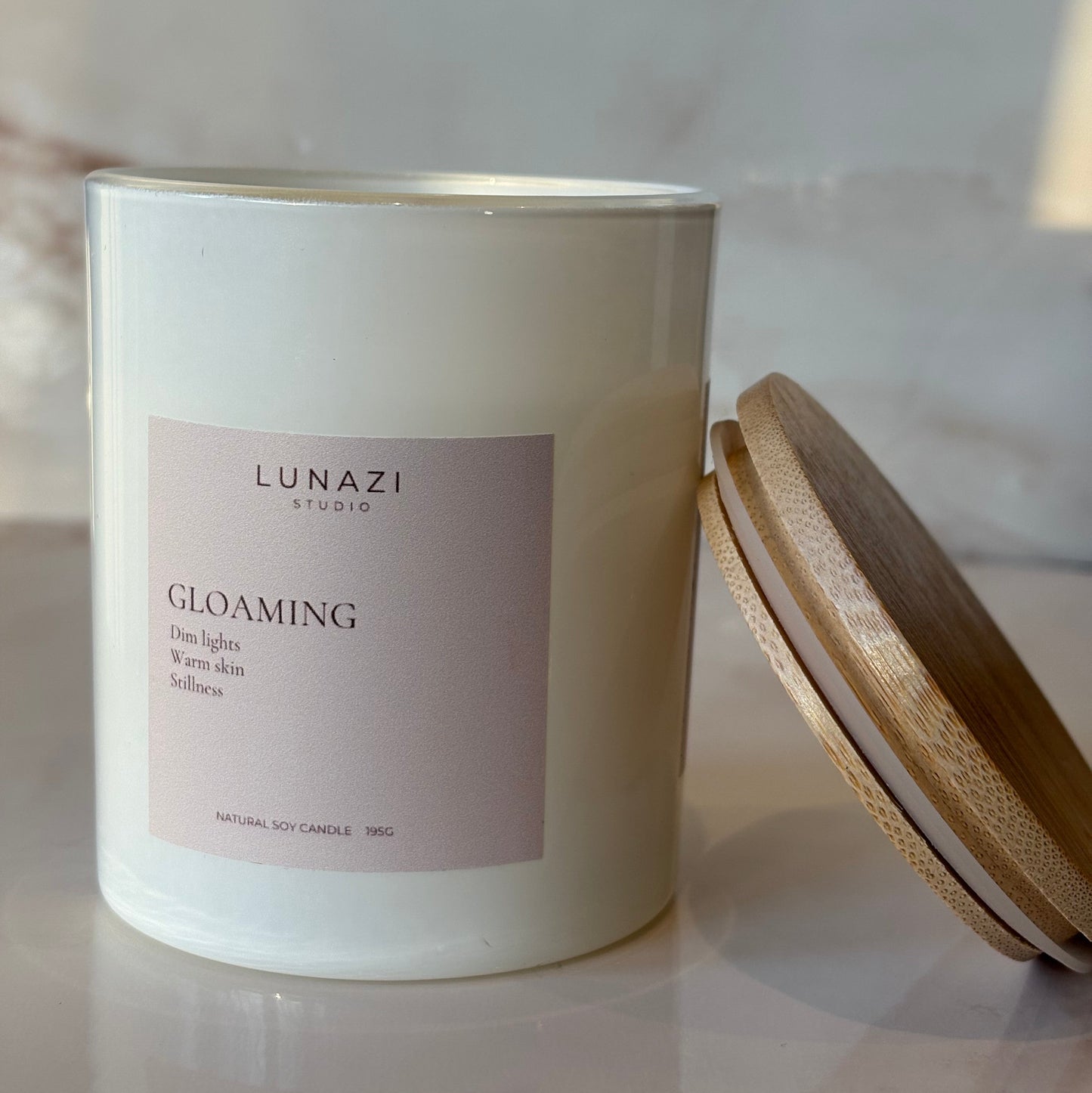 A white glass candle jar with a pale pink label reading 'LUNAZI GLOAMING' and a light wood lid resting above it on a marble-like surface.