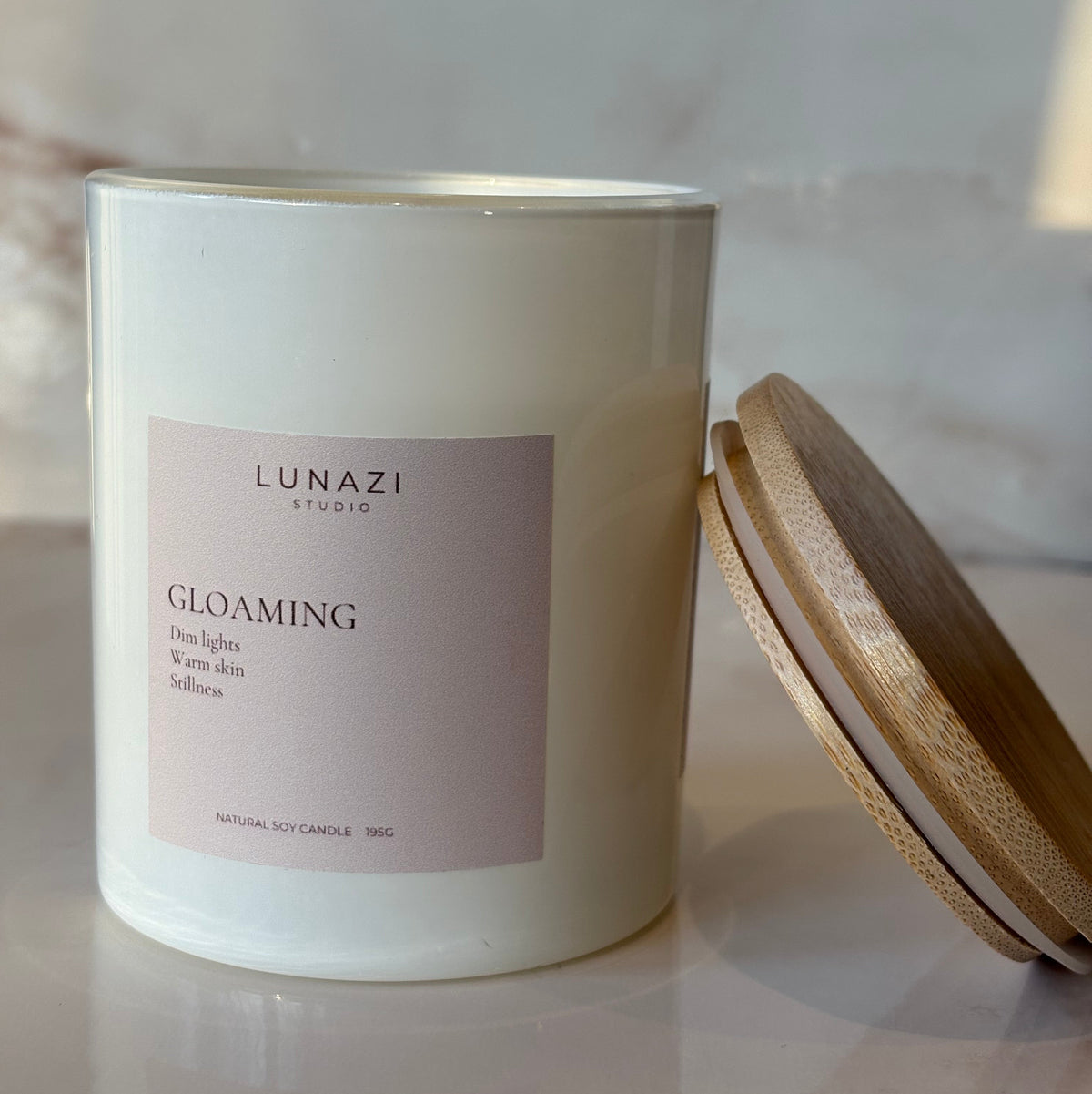 A white glass candle jar with a pale pink label reading 'LUNAZI GLOAMING' and a light wood lid resting above it on a marble-like surface.