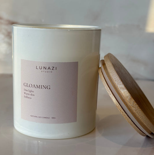 A white glass candle jar with a pale pink label reading 'LUNAZI GLOAMING' and a light wood lid resting above it on a marble-like surface.