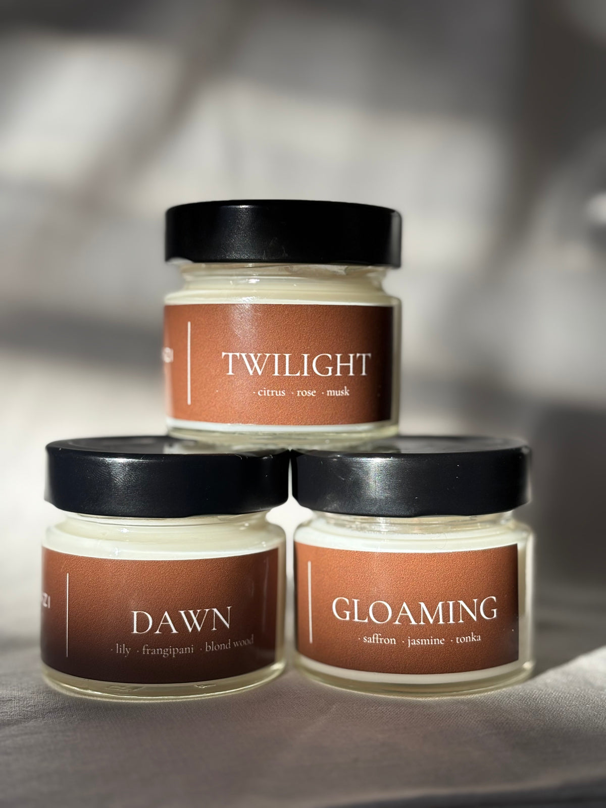 Three scented candles in clear glass jars with black lids are arranged in a pyramid. The top candle is labeled 'TWILIGHT' and the bottom two are labeled 'DAWN' and 'GLOAMING'.