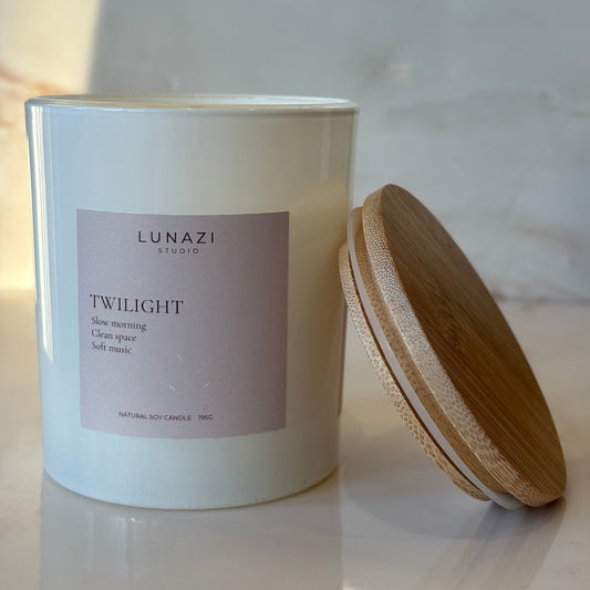 A white cylindrical candle jar with a light wood lid resting beside it on a white marble surface. The front label reads 'LUNAZI' and 'TWILIGHT'.