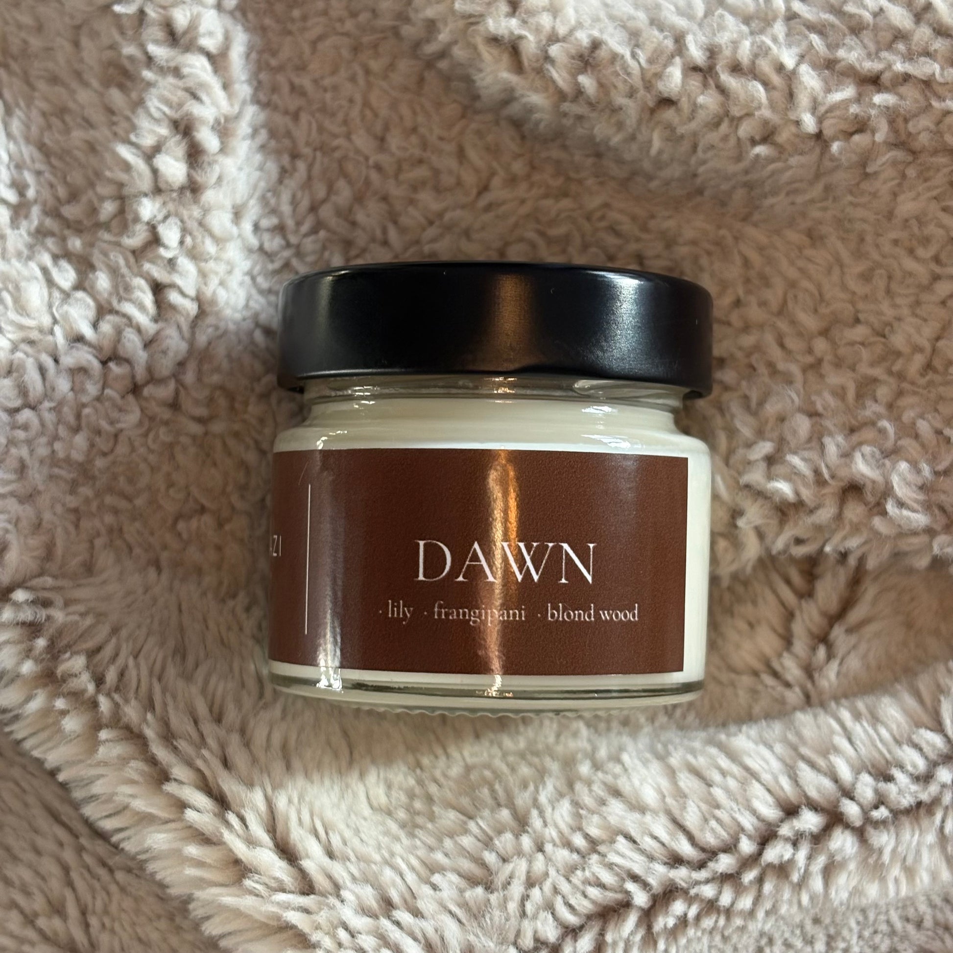 A glass jar candle with a black lid and a brown label reading 'DAWN' and 'lily - frangiani - blond wood', placed on a beige textured fleece blanket.